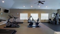 This dedicated fitness center is thoughtfully designed to provide a comfortable and functional workout environment. The space is outfitted with a selection of cardio equipment, including a treadmill, elliptical trainer, and stationary recumbent bike, all positioned to allow for easy use and movement between machines. Protective rubber floor mats are placed beneath each piece of equipment, complementing the durable, textured flooring throughout the room.

Natural light fills the space through a row of large windows fitted with white horizontal blinds, creating a bright and energizing atmosphere while offering control over light levels. Recessed ceiling lights provide additional illumination, ensuring the space remains well-lit at all times. A ceiling fan with a classic multi-blade design circulates air efficiently, keeping the environment cool and comfortable during workouts.

The walls are finished in a neutral warm tone, contributing to a clean and welcoming aesthetic. An informational posting on the wall near the entrance provides facility guidelines for users. The overall layout offers generous open floor space, allowing room for stretching or additional freestanding equipment. This fitness center is a practical and well-appointed amenity suited for a consistent and well-rounded exercise routine.