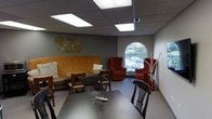 This single-floor space is designed as a comfortable and functional lounge or common area, offering a variety of seating options suited for both casual gatherings and small group meetings. The room features a built-in banquette upholstered in a warm golden fabric, accompanied by wooden dining-style chairs and tables that create a cozy, café-like corner. Complementing this arrangement, a pair of deep red armchairs adds a pop of color and a relaxed seating alternative. A standout architectural element is the arched window on the far wall, which allows an abundance of natural light to fill the room and provides a pleasant view of the surrounding outdoor greenery. The suspended drop ceiling is fitted with large recessed fluorescent light panels, ensuring consistent, even illumination throughout the space. The walls are painted in a muted gray tone, providing a neutral backdrop that highlights the warm furnishings and decorative accents, including a set of ornamental sunburst wall pieces in gold. The floor is covered in neutral gray carpeting that ties the overall palette together. The combination of varied seating, natural light, and thoughtful décor gives this space a welcoming and versatile ambiance suitable for relaxation or community use.