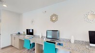 Located at 10300 University Pkwy in Pensacola, Florida, this single-floor amenity space is thoughtfully designed to serve as a productive and social hub. The interior features a clean, contemporary aesthetic with crisp white walls, light wood-style flooring, and recessed lighting throughout, creating a bright and welcoming atmosphere.

The business center area is anchored by a long granite-topped workstation with warm wood cabinetry beneath, equipped with multiple desktop computers and accented by vibrant teal ergonomic chairs. Decorative sunburst wall mirrors add a stylish touch to the workspace backdrop. A printer station is also available, making this area fully functional for everyday tasks.

Adjacent to the business center, the lounge area offers marble-topped round tables paired with modern curved chairs, ideal for casual meetings or relaxed conversation. Geometric pendant light fixtures hang overhead, complementing the overall contemporary design. Large windows along the walls allow abundant natural light to fill the space, enhancing the open and airy feel.

Further into the space, a game area with a foosball table provides a recreational outlet, while wall-mounted artwork adds color and personality throughout. The seamless flow between the work, lounge, and recreation zones makes this a versatile and well-appointed communal facility.