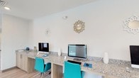 Located at 10300 University Pkwy in Pensacola, Florida, this single-floor amenity space is thoughtfully designed to serve as a productive and social hub. The interior features a clean, contemporary aesthetic with crisp white walls, light wood-style flooring, and recessed lighting throughout, creating a bright and welcoming atmosphere.

The business center area is anchored by a long granite-topped workstation with warm wood cabinetry beneath, equipped with multiple desktop computers and accented by vibrant teal ergonomic chairs. Decorative sunburst wall mirrors add a stylish touch to the workspace backdrop. A printer station is also available, making this area fully functional for everyday tasks.

Adjacent to the business center, the lounge area offers marble-topped round tables paired with modern curved chairs, ideal for casual meetings or relaxed conversation. Geometric pendant light fixtures hang overhead, complementing the overall contemporary design. Large windows along the walls allow abundant natural light to fill the space, enhancing the open and airy feel.

Further into the space, a game area with a foosball table provides a recreational outlet, while wall-mounted artwork adds color and personality throughout. The seamless flow between the work, lounge, and recreation zones makes this a versatile and well-appointed communal facility.