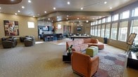 This single-floor communal space is thoughtfully designed with an open-plan layout that seamlessly connects multiple functional zones. The lounge area anchors the space with a large, L-shaped sectional sofa in warm terracotta upholstery, complemented by a matching armchair and green ottoman seating. A patterned area rug defines the lounge zone, adding texture and visual interest.

Recessed and track lighting throughout the ceiling provides consistent, well-distributed illumination across the entire space. A raised ceiling detail with angled soffits adds architectural dimension, while large windows along one wall invite abundant natural light.

A kitchen and bar area is positioned toward the rear, featuring dark cabinetry, a countertop bar with backless stools, pendant lighting in red accents, and what appears to be standard kitchen appliances. Adjacent to this is a dining area with tables and chairs, suitable for casual gatherings.

A wall-mounted flat-screen television is visible near the kitchen zone, enhancing the lounge's entertainment functionality. The flooring transitions between carpet in the seating area and hard flooring near the kitchen, clearly delineating each zone. Overall, the space presents a modern, welcoming ambiance ideal for shared social use.