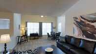 This single-story space showcases an open-concept layout that connects the living, dining, and workspace areas in one cohesive environment. The soaring vaulted ceilings create a strong sense of spaciousness and airiness, amplified by large windows dressed with horizontal blinds and flowing curtain panels that invite abundant natural light throughout the day. A ceiling fan is centrally positioned to enhance comfort and air circulation.

The living area is anchored by a dark leather sofa paired with a bold patterned area rug and a rectangular coffee table, offering a stylish and comfortable gathering space. Adjacent to this, a round dining table with upholstered chairs provides a defined yet open dining area. A dedicated workspace near the windows accommodates a desk and chair, making efficient use of the natural light available.

The flooring throughout appears to be neutral-toned carpet, contributing to a warm and inviting ambiance. The walls are finished in a soft, warm white tone that complements the overall aesthetic. The layout is thoughtfully arranged to maximize functionality while maintaining an open, uncluttered feel across the entire floor plan.