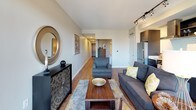 This thoughtfully designed single-floor residence offers a cohesive blend of modern finishes and functional layout throughout its open-concept floor plan. Warm hardwood-style flooring flows consistently across every room, providing visual continuity and a welcoming ambiance.

The kitchen is outfitted with sleek dark cabinetry, stainless steel appliances including a refrigerator and range, a classic white subway tile backsplash, and a wall-mounted range hood. A compact dining area positioned near a window with city views extends the kitchen's utility as an entertaining space.

The living room is bright and open, anchored by large floor-to-ceiling windows that frame urban skyline views and invite generous natural light. Track lighting overhead adds additional illumination and a contemporary touch.

The bedroom features frosted sliding doors that open to the living area, offering privacy while maintaining an airy aesthetic. Built-in closet storage is accessible directly from the bedroom.

The full bathroom includes a soaking tub with a white subway tile surround, a dark-finished vanity with a white countertop, and recessed lighting. A dedicated walk-in closet area adjacent to the bathroom provides ample organized storage with wire shelving.