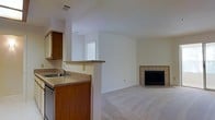 This single-floor space offers a functional and cohesive layout that connects the kitchen and living area in an open, flowing arrangement. The kitchen is equipped with warm-toned wood cabinetry, a countertop with an integrated sink, and a built-in dishwasher. A raised breakfast bar separates the kitchen from the adjoining living space, providing a natural transition between the two areas. A window above the counter allows natural light to brighten the kitchen workspace. The living area is finished with plush neutral carpeting that adds warmth and comfort underfoot. A tiled fireplace serves as the focal point of the living room, featuring a classic surround design that adds architectural character to the space. The walls are painted in a clean, neutral white throughout, creating a bright and airy atmosphere that complements the natural light filtering in. The ceiling incorporates a centrally placed ventilation duct, reflecting a practical and well-maintained interior. Overall, the space presents a straightforward, comfortable layout well-suited for everyday living, with key features such as the open-plan design, the fireplace, and the connected kitchen-living flow contributing to its functional appeal.