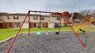 Located at Chateau Hills in Parkersburg, West Virginia, this community playground serves as a central outdoor amenity for the surrounding residential neighborhood. The play area is surfaced with a generous layer of gravel, providing a safety-conscious ground cover throughout the space.

The playground is equipped with a red metal swing set featuring multiple bucket-style toddler swings, making it suitable for young children. A yellow climbing and slide structure offers additional play opportunities, while a spring rider adds variety for smaller children. A wooden bench and waste receptacle are conveniently positioned at the edge of the play area, providing a comfortable spot for caregivers to supervise.

The playground is bordered by well-kept green lawns and is surrounded by a community of two-story brick and siding residential buildings, which feature classic red shutters and private patios. Mature trees throughout the neighborhood add seasonal color and a sense of established character. The open layout of the play area allows for clear sightlines across the space, enhancing safety and accessibility. The community setting and variety of play equipment make this playground a practical and welcoming gathering point for residents of all ages.