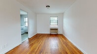 This single-floor unit offers a clean, straightforward layout with warm hardwood flooring that runs consistently throughout the living areas and bedrooms, lending a cohesive and inviting character to the space. The living room is generously proportioned, with crisp white walls, bright natural light filtering through window blinds, and an open connection to the rest of the unit. A doorway leads into a separate kitchen, which features tile flooring, brown cabinetry, a double-basin stainless steel sink with a laminate countertop, and a freestanding gas range — providing all the essentials for everyday cooking. The bedrooms are well-sized, each benefiting from natural light through windows and individual wall-mounted heating units, ensuring year-round comfort. The bathroom is finished with classic white square tile along the walls, a full soaking tub, a window for natural ventilation, and a white vanity. Throughout the unit, the clean white walls and consistent hardwood flooring create a bright, neutral canvas. The thoughtful layout, visible in the dollhouse view, provides a clear separation between living, sleeping, cooking, and bathing areas, making for a practical and functional living environment.