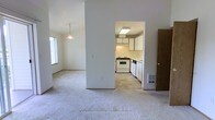 This single-floor space presents a straightforward, functional layout designed for everyday living. The main living area features wall-to-wall carpet in a neutral tone, creating a clean and understated foundation throughout. A decorative pendant light fixture hangs overhead, adding a modest focal point to the room, while a window along the side wall allows natural light to filter in, brightening the open space.

The kitchen is accessible via an open passageway from the main living area, providing a connected yet defined transition between spaces. It features white upper and lower cabinetry with light wood trim, offering ample storage. A white freestanding range and a dishwasher are included, complementing the light-toned countertops. The kitchen floor transitions to a tile-style surface, distinguishing it from the carpeted areas. A skylight or flush ceiling fixture provides additional overhead lighting within the kitchen.

A wood-paneled door is visible near the kitchen entry, likely leading to a utility or storage area. A wall-mounted floor vent is also present, indicating a centralized heating system. Overall, the space maintains a neutral, minimalist aesthetic throughout its open and connected layout.