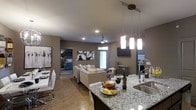 This single-floor residence is designed around an open-concept layout that seamlessly integrates the kitchen, dining, and living areas into one cohesive, flowing space. The kitchen is a standout feature, anchored by a large granite-topped island with a stainless steel undermount sink and a sleek, professional-style faucet. Stylish pendant lights hang above the island, providing focused task lighting while adding a modern decorative touch. The kitchen transitions naturally into the dining and living areas, where hardwood-style flooring runs throughout, lending warmth and continuity to the space. The living area is centered around a comfortable sectional sofa and is illuminated by recessed ceiling lighting and a ceiling fan, creating a relaxed yet functional atmosphere. A wall-mounted flat-screen television and curated wall art add personality to the living space. Neutral taupe walls provide a calming backdrop that complements the contemporary furnishings. A private bedroom is accessible directly from the main living area, offering a quiet retreat with its own distinct character. Natural light filters through windows, brightening the interior and enhancing the open, airy feel of the overall layout.