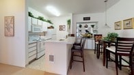 This single-floor residence offers a thoughtfully designed layout that balances comfort and functionality across its key living spaces. The entryway greets visitors with light tile flooring that transitions into plush carpet, setting a welcoming tone throughout the home. The open-concept kitchen is a practical centerpiece, featuring white cabinetry adorned with lush greenery on top, white appliances including a range, microwave, and refrigerator, and a peninsula breakfast bar with seating for multiple occupants. The kitchen flows naturally into a dining area and a cozy living room, creating an ideal space for daily living and entertaining. The bedroom is a serene retreat, carpeted for comfort and filled with natural light from a window with an exterior view. It includes a closet and ample space for furnishings. The full bathroom is clean and well-appointed, offering a long vanity with generous counter space and under-sink storage, a large wall mirror with overhead lighting, and a louvered closet door providing additional storage. Neutral wall tones and consistent finishes throughout the home create a cohesive, bright, and inviting ambiance ideal for comfortable everyday living.