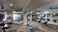 The fitness center at 1301 Rise Dr in Morrisville, NC, offers a spacious, single-floor workout facility designed to support a variety of exercise routines. The space features a distinctive checkered floor with neutral and teal-toned tiles, adding a modern aesthetic to the environment. Structural columns are integrated throughout, defining workout zones while maintaining an open, airy feel. Recessed lighting illuminates the interior, complemented by a ceiling fan that aids in air circulation during workouts. Large windows along one wall allow natural light to fill the space and provide views to the exterior. A wall-mounted flat-screen television is positioned for visibility across the cardio area, enhancing the exercise experience. The equipment layout includes a row of stationary cycling bikes with performance-style frames, along with elliptical and other cardio machines. Mirrored wall panels are incorporated into the design, visually expanding the room and allowing users to monitor their form. A metal railing along one section suggests an elevated or overlooking area adjacent to the main floor. Overall, the fitness center presents a clean, functional, and well-appointed environment suited for community use.