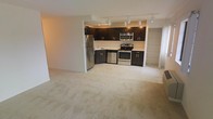 This single-floor space features an open-concept layout that seamlessly connects the kitchen and living area. The kitchen is outfitted with dark espresso-toned cabinetry paired with light countertops, creating a striking contrast. Stainless steel appliances, including a full-size refrigerator, electric range, built-in microwave, and dishwasher, provide a modern and functional cooking environment. A track lighting system mounted on the ceiling illuminates the kitchen, ensuring ample task lighting throughout the space. The kitchen transitions naturally into the expansive living area, which is finished with plush, neutral-toned carpeting that adds warmth underfoot. The open layout allows for flexible furniture arrangements and contributes to a spacious, airy feel. A partial wall separates the kitchen from the living zone, subtly defining each area while maintaining the open flow. Additional space is visible beyond the kitchen, suggesting further utility or storage areas. The overall design emphasizes practicality and a clean, contemporary aesthetic with a neutral color palette throughout.