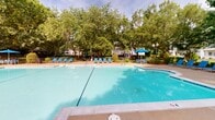 Located at 1301 Buckingham Station Dr in Midlothian, Virginia, this expansive outdoor amenity area is centered around a generously sized swimming pool with clear blue water and decorative tile detailing along the pool floor. The pool is framed by a broad concrete sun deck lined with modern blue-and-silver lounge chairs, offering ample space for sunbathing and relaxation. Umbrella-shaded seating areas provide comfortable spots to unwind out of direct sunlight. Mature trees surround the entire space, creating a naturally shaded and serene environment that enhances the sense of privacy and retreat. Carefully maintained landscaping throughout the grounds includes ornamental grasses, flowering plants, mulched garden beds, and trimmed hedgerows, all contributing to a well-kept and inviting atmosphere. A curved concrete walking path winds through the landscaped grounds, bordered by crepe myrtle trees and ornamental plantings, connecting various areas of the outdoor space. A gazebo is visible in the background, adding a charming architectural accent to the setting. Black iron fencing defines the perimeter of the amenity area, offering a sense of enclosure while maintaining open sightlines to the surrounding greenery. The overall ambiance is lush, tranquil, and resort-like.