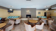 This single-floor space functions as a well-appointed communal gathering area designed to accommodate social events and relaxed lounging. The main lounge is anchored by a floor-to-ceiling gray brick fireplace, which serves as a striking focal point and is flanked by built-in bench seating with teal patterned cushions. A decorative circular wall sculpture adds an artistic touch above the fireplace, while a wall clock and wall-mounted television complete the room's functional décor. Rounded tables paired with cushioned barrel chairs are arranged throughout, creating multiple intimate seating clusters across the tile flooring.

A kitchenette area supports the space with a large laminate countertop bar, an electric range, microwave, and sink, making it suitable for catering or event preparation. Light-toned panel walls and a textured ceiling carry throughout the space.

A separate seating area opens to the outdoors through full-height sliding glass doors, offering direct views of what appears to be a pool area. This section benefits from abundant natural light and features additional table-and-chair groupings alongside a rich brown leather sofa accented with teal throw pillows, reflecting the space's consistent color palette.
