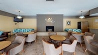 This single-floor space functions as a well-appointed communal gathering area designed to accommodate social events and relaxed lounging. The main lounge is anchored by a floor-to-ceiling gray brick fireplace, which serves as a striking focal point and is flanked by built-in bench seating with teal patterned cushions. A decorative circular wall sculpture adds an artistic touch above the fireplace, while a wall clock and wall-mounted television complete the room's functional décor. Rounded tables paired with cushioned barrel chairs are arranged throughout, creating multiple intimate seating clusters across the tile flooring.

A kitchenette area supports the space with a large laminate countertop bar, an electric range, microwave, and sink, making it suitable for catering or event preparation. Light-toned panel walls and a textured ceiling carry throughout the space.

A separate seating area opens to the outdoors through full-height sliding glass doors, offering direct views of what appears to be a pool area. This section benefits from abundant natural light and features additional table-and-chair groupings alongside a rich brown leather sofa accented with teal throw pillows, reflecting the space's consistent color palette.