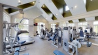 Located at 6805 West Bowles Avenue in Littleton, Colorado, this single-floor fitness center offers a thoughtfully designed workout environment suited for a wide range of exercise needs. The space is defined by its dramatic vaulted ceiling, which rises to impressive heights and is accented by striking dark geometric beam details that contrast beautifully against the crisp white surfaces. Large arched and rectangular windows line the walls, flooding the interior with natural light and offering views of the surrounding greenery, creating an open and energizing atmosphere. The flooring is finished in a neutral, durable carpet well-suited for a fitness environment. The equipment layout is spacious and well-organized, featuring a full array of cardio machines including treadmills, ellipticals, and upright bikes, as well as a variety of strength and resistance training machines positioned along the perimeter and throughout the floor. Wall-mounted televisions provide entertainment during workouts. Recessed lighting and pendant fixtures supplement the natural light, ensuring the space remains bright and functional at all hours. The overall ambiance is clean, modern, and inviting, making this fitness center a well-rounded facility for residents and users seeking a complete exercise experience.