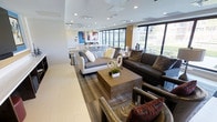 This thoughtfully designed single-floor amenity space offers a seamless blend of comfort and contemporary style. The open-plan layout is divided into distinct yet connected zones, creating a versatile environment suited for both relaxed socializing and casual gatherings.

The main lounge area features a curated mix of seating, including a plush white sofa, a rich dark leather couch, and accent chairs with vibrant patterned cushions, all centered around a solid wood coffee table. Wide-plank wood-look flooring anchors the space and transitions smoothly into light tile near the perimeter.

A dedicated media zone is framed by a bold blue accent wall, housing a flat-screen television flanked by contemporary artwork and built-in cabinetry offering generous storage. Adjacent to this area, open shelving and a tiled backsplash accent a beverage and prep station equipped with small appliances, adding a functional hospitality element.

A striking marble-top communal table with sleek acrylic chairs serves as both a dining and collaboration area, positioned near a row of floor-to-ceiling windows that flood the interior with natural light and provide open views of the surrounding greenery. Recessed lighting throughout ensures a warm, well-lit ambiance at any hour, completing this polished and welcoming communal retreat.