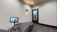 This single-room wellness and recovery space is thoughtfully designed to provide a tranquil retreat within a fitness environment. The room features a contoured ergonomic relaxation lounger as its centerpiece, upholstered in sleek dark leather and positioned to maximize comfort during rest or recovery sessions. A mounted display screen is stationed beside the lounger, offering interactive or guided wellness content for users during their session.

The interior design strikes a balance between modern and serene. Neutral light grey walls are paired with dark baseboard trim and textured grey carpeting, creating a grounded, calming atmosphere. Warm, directional wall sconces with a contemporary black and gold finish provide soft, ambient lighting throughout the room.

A standout design element is the large-format zen-inspired wall mural depicting stacked meditation stones set against a sandy backdrop, anchoring one corner of the room with a sense of mindfulness and calm. Adjacent to it, a subtle wood-textured accent wall adds visual depth and warmth.

A glass-paneled black door connects the recovery room directly to the main fitness floor, visible through the pane and equipped with smart control panels beside the entry. Recessed ceiling lighting supplements the ambient wall sconces, ensuring a well-lit yet relaxing environment. The overall ambiance is polished, purposeful, and conducive to post-workout recovery or guided relaxation.