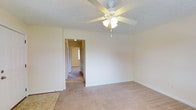 This single-story space presents a clean, functional layout with a warm and understated aesthetic. The main room is finished with wall-to-wall carpet in a neutral beige tone, complemented by smooth, cream-colored walls that create a bright and open atmosphere. A white five-blade ceiling fan with an integrated light fixture serves as the room's central overhead element, providing both ventilation and ambient lighting.

A doorway leads into a connecting hallway, which provides access to additional rooms throughout the space. The hallway is similarly carpeted and features multiple doors along its length, suggesting several private rooms branching off from this central corridor. A small window at the far end of the hallway allows natural light to filter through, adding depth and brightness to the interior passage.

The textured ceiling and clean white baseboards contribute to a tidy, well-maintained appearance throughout. The overall design is straightforward and practical, with a neutral color palette that lends itself well to a variety of furnishing styles. The space offers a comfortable, move-in-ready environment suitable for everyday living.