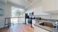 This well-designed single-floor unit offers a clean, contemporary aesthetic with a thoughtfully arranged open-concept layout. Light wood-look flooring flows consistently throughout, tying each area together seamlessly.

The kitchen is a standout feature, outfitted with sleek white cabinetry extending to the ceiling, neutral gray countertops, and a full suite of stainless steel appliances including an electric range, over-the-range microwave, refrigerator, and dishwasher. A large sliding glass door with vertical blinds fills the kitchen with natural light and offers views of lush palm trees beyond the balcony.

The living area is bright and airy, benefiting from additional floor-to-ceiling sliding glass doors that invite natural light deep into the space. A wall-mounted media unit with open shelving serves as a clean focal point, complemented by neutral walls and understated decor.

The bedroom is a calm retreat, featuring a generously sized window dressed with vertical blinds that allow for adjustable natural light and outdoor views. The soft, neutral palette continues here, creating a restful atmosphere.

The bathroom is finished with classic white tile flooring, a marble-topped vanity, and a tub-shower combination, maintaining the unit's clean and functional design language throughout.