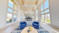 This expansive game room is a well-appointed community amenity space designed for both recreation and relaxation. The interior is defined by dramatically high ceilings with coffered detailing and a series of elegant geometric pendant chandeliers that lend a refined yet inviting atmosphere. Light hardwood-style flooring runs throughout, complementing the crisp white walls and bright, airy aesthetic.

Floor-to-ceiling windows line the perimeter, flooding the space with abundant natural light and offering unobstructed views of the surrounding landscaped grounds. The entryway features a decorative tile inlay and glass-paneled double doors, creating a welcoming first impression. A striking marble-finished accent wall anchors one side of the space near the entrance.

The main living area includes a comfortable lounge zone with bold blue upholstered seating arranged around a central coffee table, anchored by a patterned area rug. A full-size pool table serves as the recreational centerpiece, with additional seating and game tables nearby for versatile use.

The kitchen is outfitted with stainless steel appliances, granite countertops, gray cabinetry, and a vivid ocean-blue fish-scale mosaic tile backsplash that adds a coastal, artistic flair. A large island provides additional prep and gathering space. Together, these elements create a dynamic, multi-functional environment ideal for social gatherings and leisure activities.