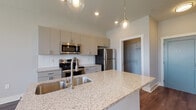 Located at 2875 Olentangy River Rd in Columbus, Ohio, this single-floor residence offers a clean, contemporary design with thoughtful finishes throughout. The kitchen serves as a central highlight, featuring a large granite-topped island with an undermount stainless steel sink, pendant globe lighting, and a full suite of stainless steel appliances including a refrigerator, range, and microwave. Flat-panel light gray cabinetry with sleek hardware lines the walls, complemented by a white subway tile backsplash and generous counter space. An open layout connects the kitchen to the living area, all unified by durable wood-style plank flooring in a warm gray tone. The living space benefits from abundant natural light streaming through multiple large windows fitted with horizontal blinds. The bathroom is finished consistently with the rest of the unit, featuring a granite-topped vanity, gray cabinetry, and a mirror with a marble-patterned frame, alongside a combined tub and shower with frosted glass doors. A dedicated closet with built-in shelving and hanging rods provides organized storage. The overall aesthetic is modern and neutral, offering a bright, move-in-ready environment with a cohesive design palette throughout.