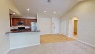 This single-floor unit is defined by its striking vaulted ceilings in the main living area, which rise dramatically and are complemented by recessed lighting and a skylight that floods the space with natural light. The open-concept layout connects the living room seamlessly to the kitchen, with transitions between plush carpeting in the living area and light hardwood-style flooring in the kitchen and entryway zones.

The kitchen is well-appointed with rich medium-toned wood cabinetry, dark granite countertops, and stainless steel appliances including a refrigerator, stove, and microwave. A peninsula counter serves as a natural divider between the kitchen and living space. The front entry is accessible via a paneled white door, with an arched passageway leading further into the unit.

The bedrooms are carpeted and finished in warm neutral tones, each offering windows with exterior views and ample space. Two bathrooms are present — one featuring a vanity with wood cabinetry, a wall mirror, and a shower-tub combination, and a second with a tub, toilet, and grab bars. Both bathrooms are finished with light hardwood-style flooring and warm beige walls, maintaining a consistent aesthetic throughout the unit.