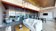 This single-floor amenity space is designed with a polished, contemporary aesthetic that balances elegance with functionality. The main lounge area is anchored by a large cream sofa and complemented by light grey upholstered seating arrangements, all resting on patterned carpet that adds texture to the refined interior. Striking Sputnik-style chandeliers with globe bulbs hang from the ceiling, casting warm ambient light across the space. A bold red ceiling panel contrasts with gold-toned accents, creating a visually dynamic overhead detail that defines different zones within the lounge.

Floor-to-ceiling glass partitions divide the space into distinct seating areas while maintaining an open, airy feel and allowing natural light to flow throughout. A formal dining area features a long dark table with seating for several guests, positioned directly in front of expansive windows that frame sweeping city skyline views. High-top bar seating lines the perimeter near the windows, offering additional vantage points overlooking the cityscape.

A dedicated workspace section provides individual desks with city-facing windows, shelving, and built-in surfaces suited for focused work. Decorative gold disc wall art and curated accessories throughout reinforce the space's upscale, design-forward character.