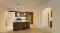This single-floor residence showcases a thoughtfully designed open-concept layout that connects the kitchen and living areas in a warm, cohesive fashion. The kitchen is a standout feature, offering rich dark-stained wood cabinetry that extends to the ceiling for generous storage capacity. Stainless steel appliances, including a refrigerator, gas range, and built-in microwave, provide a modern, functional edge. A curved kitchen island with a granite countertop and an integrated sink serves as both a practical workspace and a natural gathering point. Pendant lighting hangs above the island, adding a refined touch, while recessed track lighting and decorative ceiling spotlights illuminate the space throughout. A classic tile backsplash runs along the cooking area, adding texture and visual interest. Crown molding accents the ceiling, lending an elegant architectural detail. An arched entryway gracefully transitions from the kitchen into the adjacent carpeted living area, enhancing the open and airy feel of the home. Neutral tones in the walls and flooring create a versatile backdrop, making the space feel inviting and easy to personalize.