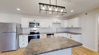 This contemporary single-floor unit offers a thoughtfully designed open-concept layout, where the kitchen flows seamlessly into the expansive living area. The kitchen is a standout feature, showcasing crisp white shaker cabinets with matte black hardware, a striking herringbone tile backsplash, and a generous center island topped with rich granite-style countertops. A bold industrial-style pendant light anchors the island, while recessed lighting throughout ensures the space is well-lit. Stainless steel appliances, including a refrigerator, range, microwave, and dishwasher, complete the kitchen's modern aesthetic. Light hardwood flooring runs continuously through the main living areas and bedrooms, creating a warm and cohesive feel. The bedrooms are bright and airy, with natural light filtering through windows fitted with blinds. Closet space is provided with built-in shelving and hanging rods. A full bathroom features a tub-shower combination and neutral tile flooring. The dedicated laundry area includes a full-size washer and dryer. A private balcony with metal railing offers views of the surrounding neighborhood and tree-lined street. The overall design is clean, modern, and move-in ready.
