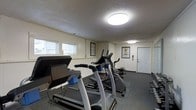 This fitness center offers a practical and well-organized workout space designed to accommodate a variety of exercise needs. The room features a textured ceiling with flush-mount circular light fixtures that provide bright, even illumination throughout the space. A row of windows along one wall allows natural light to filter in, complementing the overhead lighting and contributing to an open, energizing atmosphere.

The flooring is covered in durable dark carpeting, well-suited for a high-traffic fitness environment. The space is equipped with multiple cardio machines, including treadmills and an elliptical trainer, arranged in a row to maximize the available floor area. Additional strength and conditioning equipment is visible toward the rear of the room, offering users a range of workout options.

Motivational and instructional fitness posters are mounted on the walls, providing helpful exercise guidance. A fire safety extinguisher is also visibly mounted near the exit door, reflecting attention to safety standards. The single entry door leads conveniently in and out of the space. Overall, this fitness center presents a clean, functional environment with everything needed for an effective cardio and strength training session.