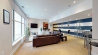 This well-appointed single-floor space is designed for comfort and social gathering, offering an open-concept layout that seamlessly connects multiple functional zones. The main lounge area is anchored by rich dark brown leather sofas arranged around glass-topped coffee tables, providing ample seating for groups. A striking navy blue and white horizontal striped accent wall serves as a bold focal point, adorned with framed artwork and complemented by high bar-height tables and stools — ideal for casual conversation or informal dining.

Recessed lighting throughout the crisp white ceiling creates a bright, inviting atmosphere, while oversized floor-to-ceiling windows along one wall flood the space with natural light and offer views of the surrounding streetscape. A wall-mounted flat-screen television enhances the lounge's entertainment appeal.

Tucked within the space is a fully equipped kitchenette featuring warm cherry wood cabinetry, dark granite countertops, tile flooring, and a complete suite of stainless steel appliances including a refrigerator, oven, microwave, and dishwasher — making it well-suited for catered events or everyday use. A city skyline silhouette mural adds an urban artistic touch to one of the accent walls, reinforcing the sophisticated, contemporary character of the overall space.