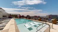 Located atop a high-rise building at 8621 Georgia Ave in Silver Spring, Maryland, this rooftop pool deck offers a striking urban retreat with expansive open-air amenities and commanding views of the surrounding cityscape. The centerpiece of the space is a generously sized swimming pool with crystal-clear turquoise water, in-pool lounge chairs for relaxing at the water's edge, and accessible entry features including a pool lift. The pool deck is finished with large-format stone-toned pavers that provide a clean, modern aesthetic throughout. Surrounding the pool, a broad sun deck is furnished with rows of sling-style lounge chairs, providing ample space for sunbathing and outdoor lounging. Additional seating areas include cushioned pod chairs and shaded spots beneath a red umbrella, catering to a variety of relaxation preferences. Planters with seasonal flowers and greenery add a touch of nature to the elevated setting. The deck is partially framed by a warm brick masonry wall with horizontal windows that reflect the building's architectural character, while the opposite side opens to sweeping panoramic views of the Silver Spring and greater Washington, D.C. metro skyline. A glass-enclosed indoor amenity room is accessible directly from the pool deck, seamlessly connecting indoor and outdoor spaces.