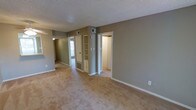 This single-story space features a straightforward, functional layout with a neutral color palette throughout. The main living area is finished with wall-to-wall carpeting in a warm beige tone, complemented by grey and taupe painted walls that create a calm, understated ambiance. A textured popcorn ceiling adds a classic touch overhead, while white baseboards provide clean visual contrast along the floor line.

A built-in shelving unit with a cabinet base is integrated into one wall, offering practical display and storage space. Multiple HVAC vents are visible both on the walls and floor, ensuring climate control throughout. Wide doorways connect the main area to adjacent rooms, one of which appears to be a hallway or corridor leading toward a windowed entry door, allowing natural light to filter into the interior. An additional open doorway leads to a brightly lit adjoining room, suggesting a bedroom or utility space.

The overall layout flows openly from room to room, with consistent flooring and finishes tying the spaces together. The design is practical and move-in ready, providing a clean, neutral canvas well-suited for a variety of interior arrangements.