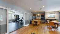 Located at 1524 Lakeview Dr in Darien, IL, this single-floor amenity space is designed to serve as a well-rounded gathering and wellness facility. The layout integrates several distinct functional areas that flow cohesively together, unified by consistent soft blue-gray wall tones, white chair-rail molding, and warm hardwood flooring throughout the common areas.

The fitness center occupies a generously sized room with durable rubber flooring and is well-lit by multiple windows that invite natural light and offer views of the surrounding greenery. The space is equipped with cardio machines including an elliptical, recumbent bike, and treadmill, as well as a multi-station cable machine and a dumbbell rack. A wall-mounted television and ceiling fan add to the practicality of the room, and vaulted ceiling details lend it an open, airy character. Frosted glass panel doors connect the fitness area to the adjacent common spaces, maintaining visual separation while preserving an open feel.

The kitchen area features stainless steel appliances, including a refrigerator, electric range, and over-range microwave, complemented by warm wood cabinetry. A breakfast bar with seating and a pass-through window adds casual dining functionality alongside a round dining table with seating for four.

The lounge area provides a relaxed retreat anchored by a dramatic floor-to-ceiling fieldstone fireplace, with comfortable seating, hardwood floors, and recessed lighting completing the inviting atmosphere.