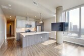 Located on the 18th floor at 888 Bellevue, this two-bedroom, two-bathroom residence offers a thoughtfully designed open-concept layout with contemporary finishes throughout. The combined kitchen and living area serves as the heart of the home, showcasing a large central island with a white quartz countertop, two-tone cabinetry in glossy white and warm wood tones, stainless steel appliances, and stylish pendant lighting. Exposed concrete columns add an industrial architectural accent that contrasts beautifully with the light hardwood-style flooring. Floor-to-ceiling windows flood the living space with natural light and frame sweeping views of the downtown Bellevue skyline. A hallway connects the main living area to both bedrooms, each offering generous proportions, plush carpet, and large windows with dramatic city views. The spacious primary bedroom features its own en-suite bathroom equipped with a floating dark wood vanity, a backlit mirror, and wood-look flooring. The second bathroom includes a soaking tub with marble-effect tile surround and wood-tone cabinetry. Three closets provide ample storage throughout. The unit's clean lines, neutral palette, and high-end finishes create a polished, modern ambiance ideal for urban living.
