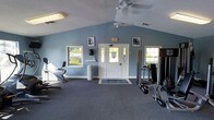 This single-story fitness room offers a welcoming and functional workout environment highlighted by a vaulted ceiling that adds a sense of openness and airiness to the space. The walls are painted in a calming blue-gray tone, contributing to a clean and motivating atmosphere. A ceiling fan is centrally mounted, providing consistent air circulation throughout the room.

Natural light filters in through several windows positioned along the walls, offering views of surrounding greenery and enhancing the overall brightness of the space. The carpeted flooring provides a comfortable and stable surface underfoot. The room is equipped with a selection of fitness equipment, including cardio machines and a multi-use strength training station with weight attachments, catering to a variety of workout routines.

A wall-mounted mirror on one side allows users to monitor their form during exercise. Framed motivational artwork decorates the walls, adding a purposeful aesthetic touch. The centrally located entry door features sidelights that further contribute to the natural light. A textured ceiling and clearly marked emergency exit signage complete the practical, well-appointed design of this fitness space.