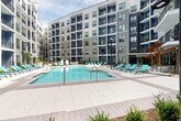 This outdoor amenity space is set within a contemporary multi-story building complex defined by a bold architectural palette of navy blue, gray, and white paneling with large windows and private balconies framing the courtyard. At the center of the space is a generously sized swimming pool with clear blue water, surrounded by an expansive pool deck furnished with aqua-colored lounge chairs arranged for optimal relaxation and sunbathing.

Adjacent to the pool, a built-in outdoor kitchen station features brick construction, marble-style countertops, and dual stainless steel grills, providing a well-equipped space for outdoor cooking and entertaining. Nearby, a covered lounge area offers shade and comfort, distinguished by a wood-slat ceiling, bold geometric patterned tile accent walls in terracotta, black, and white, and an assortment of seating including blue sectional sofas and accent chairs. The mixed-material flooring combines large-format pavers and wood-look decking, adding texture and visual interest throughout. Landscaping elements including ornamental shrubs and trees soften the hardscape, contributing to an inviting, resort-like ambiance within the courtyard.