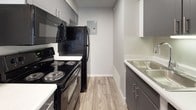 This single-floor unit offers a clean, contemporary design with a neutral gray color palette throughout. The galley-style kitchen is efficiently laid out and features gray cabinetry, white countertops, a stainless steel sink, and a full suite of black appliances including a refrigerator, electric range, and over-the-range microwave. Wood-look vinyl plank flooring runs throughout the kitchen and living areas, providing a cohesive and easy-to-maintain surface. A structural column subtly defines the transition between the kitchen and the adjacent living space, which is open and airy with a ceiling-mounted light fixture.

The living area is bright and versatile, with light wood-look flooring and a large window that invites natural light. The textured ceiling adds a layer of architectural character shared throughout the unit. The bedroom is a comfortable, well-proportioned space finished with soft, neutral carpeting and its own window fitted with blinds, allowing for adjustable natural light and privacy. Clean white baseboards and consistent gray wall tones create a harmonious, move-in-ready aesthetic. The hallway connects the rooms efficiently, with white interior doors completing the polished, modern feel of the space.