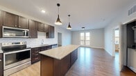 Located at 1011 Lusso Ave in Columbus, Ohio, this single-floor residence offers a thoughtfully designed open-concept layout finished with consistent light wood-tone flooring throughout. The kitchen serves as a central focal point, showcasing rich dark-brown cabinetry that extends to the ceiling for generous storage, a classic white subway tile backsplash, and a sizeable granite-topped island illuminated by stylish black pendant lights. Stainless steel appliances, including a French door refrigerator, range, microwave, and dishwasher, complete the functional and polished kitchen space. The kitchen flows seamlessly into a spacious living area bathed in natural light, with French doors opening to a private balcony overlooking the outdoors. The bedrooms are well-proportioned, featuring neutral walls and ample walk-in closet space outfitted with wire shelving systems. The primary bathroom is notably spacious, offering a dual-sink vanity with dark cabinetry and a white countertop, a large mirror with overhead lighting, and a glass-enclosed shower. A secondary bathroom echoes the same refined design language with a single-sink vanity and bright, clean finishes. The overall ambiance is modern, clean, and cohesive.