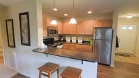 This single-floor residence offers a thoughtfully designed layout centered around a functional and stylish kitchen. The kitchen is a standout feature, showcasing warm honey-toned wood cabinetry that extends to the upper walls, providing generous storage throughout. Dark granite countertops add a rich contrast, running along the workspace and wrapping around a curved peninsula that doubles as a breakfast bar, complete with wooden bar stools for casual dining. Stainless steel appliances, including a refrigerator and microwave, lend a modern, polished look, while a black range adds a bold accent. Two elegant pendant lights hang above the peninsula, casting warm, focused light over the bar area and complementing the recessed lighting overhead. The kitchen opens naturally to the adjoining living space, creating a connected, open-concept feel that encourages easy movement throughout the home. Light-toned walls and wood-style flooring contribute to a bright, welcoming ambiance. Overall, the space strikes a balance between practicality and comfort, making it well-suited for both everyday living and entertaining.
