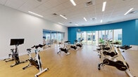 Located at 1517 Cameron Samuel Ln in West Palm Beach, Florida, this single-floor fitness facility offers a well-designed, energetic environment for both exercise and recreation. The space is divided into two distinct areas, each thoughtfully appointed to serve its purpose.

The main fitness studio is a generously sized spin room featuring rows of Spinning bikes arranged for group cycling classes. Bold teal accent walls contrast with crisp white surfaces, creating a dynamic and motivating atmosphere. Light wood-style flooring runs throughout, while a drop-tile ceiling with recessed linear LED lighting ensures bright, even illumination. Large floor-to-ceiling glass doors and windows along one wall provide abundant natural light and direct views of lush tropical landscaping outside. Wall-mounted flat-screen TVs and a full-length mirror enhance the workout experience, and a compact equipment area with dumbbells and accessories adds additional functionality.

The second room serves as a children's play area, featuring the same warm wood flooring and a cheerful blue ceiling. This welcoming space includes a wooden climbing structure with a slide, a chalkboard panel, open shelving stocked with toys and activities, and ride-on toys scattered across the open floor. A large mirror connects it visually to the fitness area, and windows overlooking tropical greenery fill the room with natural light.