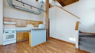 This two-story space showcases a striking industrial-meets-rustic aesthetic defined by its dramatic vaulted ceilings with exposed wooden beams and visible ductwork. The upper level is accessible via a staircase visible from the main floor, adding vertical dimension and a sense of spaciousness to the interior.

The kitchen area is outfitted with warm honey-toned oak cabinetry, a peninsula with a light-colored countertop, and a sink. A compact appliance station is integrated alongside the cabinetry, and track lighting mounted to the ceiling provides focused illumination throughout the open space.

The main living area features light hardwood flooring that runs throughout, complementing the natural wood tones of the structural beams and cabinetry. The soaring ceiling height draws the eye upward, where exposed metal ductwork and wooden ceiling panels reinforce the industrial character of the space. Structural wood posts punctuate the open floor plan, adding architectural interest.

Natural light filters in, highlighting the clean white walls that contrast with the rich wood elements. The open layout between the kitchen and living areas promotes a flexible, loft-like atmosphere throughout.