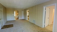 This single-floor home presents a practical and functional layout designed for comfortable everyday living. The main living area is generously sized, finished with wall-to-wall carpeting that adds a sense of warmth underfoot. Neutral, light beige walls throughout create a clean and versatile backdrop, making the space easy to personalize. A floor vent indicates a forced-air climate system, ensuring year-round comfort.

From the living area, an open passageway leads directly into the kitchen, which is visible in the background and features standard appliances and cabinetry. Natural light filters in through a window in the kitchen, brightening the adjacent spaces. A second doorway from the living area provides access to an additional room, also receiving natural light from its own window, suggesting it may serve as a bedroom or secondary living space.

A large closet or utility space is accessible via a set of double doors along the interior wall, offering convenient built-in storage. The textured ceiling adds subtle architectural character to the otherwise clean and understated interior. Overall, the home offers a no-frills, move-in-ready layout with a cohesive neutral palette and practical room connectivity throughout its single level.