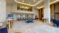 This elegantly designed amenity lounge makes an immediate impression with its soaring double-height ceilings adorned with an intricate layered tray design and warm ambient lighting. The grand entrance welcomes guests through black-framed glass double doors flanked by sheer floor-to-ceiling drapes, with herringbone-patterned hardwood flooring flowing throughout the space.

The main gathering area centers around a sleek billiards table set atop a large area rug, creating a natural focal point within the expansive open layout. Adjacent to it, a curved wet bar with a dark granite countertop and wood-paneled base is equipped with bar seating and dual mounted flat-screen televisions, making it ideal for social gatherings. Pendant lights with gold accents hang above the bar, adding a warm, refined touch.

Flanking the main space are intimate seating areas, including a generous sectional sofa positioned beside floor-to-ceiling windows that offer views of the surrounding streetscape. Striking veined marble wall panels serve as dramatic design elements throughout.

A private conference-style dining room accommodates a full-length wooden table with upholstered gold-framed chairs, a wall-mounted television, and tall black-framed windows that fill the room with natural light. An upper mezzanine level overlooks the lounge below, enhancing the dramatic vertical scale of this upscale communal space.
