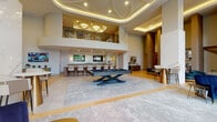 This elegantly designed amenity lounge makes an immediate impression with its soaring double-height ceilings adorned with an intricate layered tray design and warm ambient lighting. The grand entrance welcomes guests through black-framed glass double doors flanked by sheer floor-to-ceiling drapes, with herringbone-patterned hardwood flooring flowing throughout the space.

The main gathering area centers around a sleek billiards table set atop a large area rug, creating a natural focal point within the expansive open layout. Adjacent to it, a curved wet bar with a dark granite countertop and wood-paneled base is equipped with bar seating and dual mounted flat-screen televisions, making it ideal for social gatherings. Pendant lights with gold accents hang above the bar, adding a warm, refined touch.

Flanking the main space are intimate seating areas, including a generous sectional sofa positioned beside floor-to-ceiling windows that offer views of the surrounding streetscape. Striking veined marble wall panels serve as dramatic design elements throughout.

A private conference-style dining room accommodates a full-length wooden table with upholstered gold-framed chairs, a wall-mounted television, and tall black-framed windows that fill the room with natural light. An upper mezzanine level overlooks the lounge below, enhancing the dramatic vertical scale of this upscale communal space.