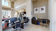 Located on Harvey Road in College Station, Texas, this single-story community clubhouse and business center offers a polished, functional environment designed to support both productivity and leisure. The space is characterized by soaring ceilings that amplify the sense of openness throughout, with large windows and glass-paneled entry doors allowing abundant natural light to fill the interior. Sheer white curtains frame the windows, offering views of the landscaped exterior grounds.

The business center area is equipped with multiple desktop computer workstations arranged along dark wood-toned desks, each paired with ergonomic rolling chairs. A dedicated printer and supply station sits along one wall, complemented by built-in cabinetry for storage. Contemporary abstract artwork decorates the warm taupe walls, contributing to a professional yet welcoming atmosphere.

Light-toned tile flooring extends throughout the space, connecting the business center seamlessly to a wide hallway entry corridor and adjoining common areas. Beyond the business center, the open layout reveals a well-appointed lounge area and a game room featuring a billiards table, along with a kitchen counter space with pendant lighting. The overall design blends practicality with upscale residential aesthetics, creating a cohesive and comfortable shared environment.