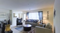 Located at 53 Sand Pit Rd in Danbury, Connecticut, this single-floor residence offers a welcoming and thoughtfully designed living environment. The open-concept layout creates a seamless flow between the living and dining areas, making the space feel expansive and airy.

The living room is a standout feature, bathed in natural light streaming through large windows dressed with flowing blue curtains. Neutral-toned walls and light carpeting provide a clean, versatile backdrop, complemented by stylish furnishings including a plush sofa adorned with decorative pillows, a pair of armchairs, and a round leather ottoman that serves as a functional centerpiece. Warm accent lighting from table lamps adds a cozy ambiance throughout.

Adjacent to the living area, the dining space is visible through an open pass-through, featuring track lighting overhead and a well-appointed dining set, reinforcing the home's cohesive and functional design. The overall atmosphere is bright, calm, and inviting, with a tasteful blend of contemporary and classic design elements that create a comfortable and livable environment suitable for everyday living and entertaining alike.
