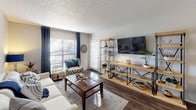 This single-story residence offers a thoughtfully designed layout that balances comfort and functionality across its main living spaces. The living room serves as the heart of the home, featuring dark hardwood-style flooring, a textured ceiling, and a large window dressed with navy drape panels that allow ample natural light to fill the room. A sliding door provides additional access to the outdoors.

The dining area is distinguished by a bold navy accent wall that creates a striking focal point, complemented by a stylish pendant light overhead and a pass-through opening connecting directly to the kitchen. The kitchen is laid out in a galley configuration, equipped with white cabinetry, subway tile backsplash, light stone countertops, a stainless steel sink, and black appliances, including an electric range.

Two bedrooms provide comfortable retreats; one features a teal accent wall with soft carpeting, while the second is finished in neutral tones with a window overlooking the outdoors. The full bathroom includes a tub-shower combination with tile surround, a single-sink vanity with stone countertop, a large mirror, and wood-style flooring, presenting a clean and practical design throughout.