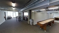This single-floor common area combines an industrial aesthetic with casual comfort, creating a functional space designed for social interaction and leisure. The ceiling features exposed metallic ductwork and conduit running horizontally across a dark-painted surface, lending the space a contemporary, industrial character. Fluorescent lighting is integrated throughout, ensuring consistent illumination across the entire area.

The layout is divided into distinct zones. A seating area near the windows includes a sofa and additional soft furnishings, providing a relaxed lounge environment. Natural light filters in through several windows, brightening that section of the space. Nearby, a set of high-top tables and bar-height chairs offers a more upright seating option, suitable for casual conversation or light work.

A shuffleboard table positioned toward the perimeter adds a recreational element, signaling that the space is intended for entertainment and community use. Structural columns punctuate the open floor plan, subtly defining zones without fully enclosing them. The carpet flooring in a neutral tone ties the areas together cohesively. Overall, the space balances functional amenities with an approachable, community-oriented atmosphere.