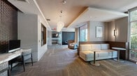 Located at 40 Pinehills Drive in Plymouth, Massachusetts, this single-floor communal space offers a refined and welcoming environment designed for both relaxation and productivity. The open lounge area is anchored by stylish globe pendant chandeliers that hang from a coffered ceiling, casting a warm glow over plush white sofas, accent chairs, and contemporary side tables arranged atop patterned carpeting in neutral tones. Warm taupe and gray walls are accented by framed artwork, decorative wall panels, and subtle sconce lighting that contribute to the space's polished ambiance. Floor-to-ceiling windows in the seating nook flood the interior with natural light while offering lush, wooded views of the surrounding Pinehills landscape — a serene backdrop that enhances the overall atmosphere. A dedicated conference room features a sleek, white rectangular table surrounded by ergonomic chairs, a wall-mounted flat-screen display set against a textured feature wall, and a large whiteboard, making it well-suited for meetings and presentations. A small reading nook with a bookshelf and armchair adds a casual, community-oriented touch. Throughout, the design blends modern aesthetics with comfortable, functional furnishings to create a cohesive and inviting space.